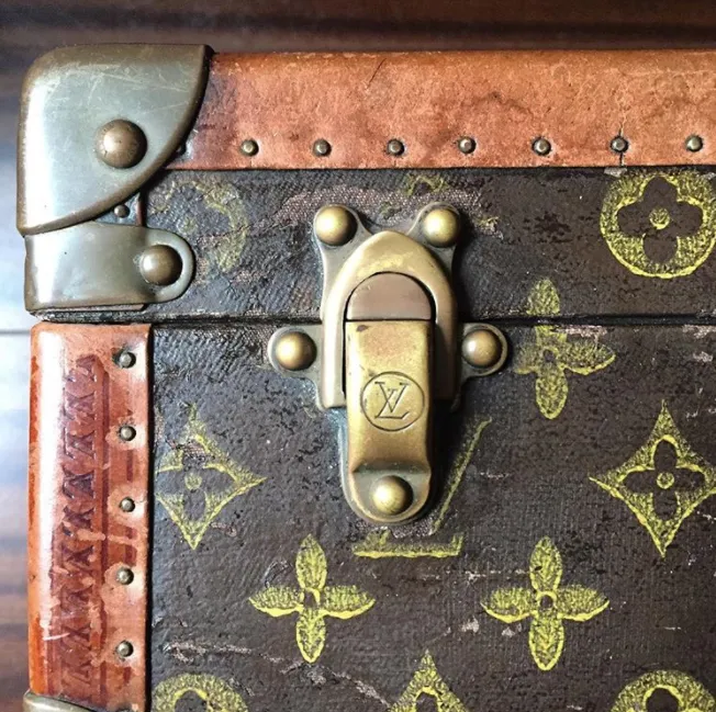 A close-up of a vintage Louis Vuitton trunk featuring the brand’s iconic monogram canvas with floral and LV logo patterns. The trunk showcases aged leather trim with metal reinforcements, brass studs, and a brass lock embossed with the LV initials. The wear on the leather and metal gives it an antique, well-traveled look, highlighting Louis Vuitton's legacy in luxury luggage craftsmanship.