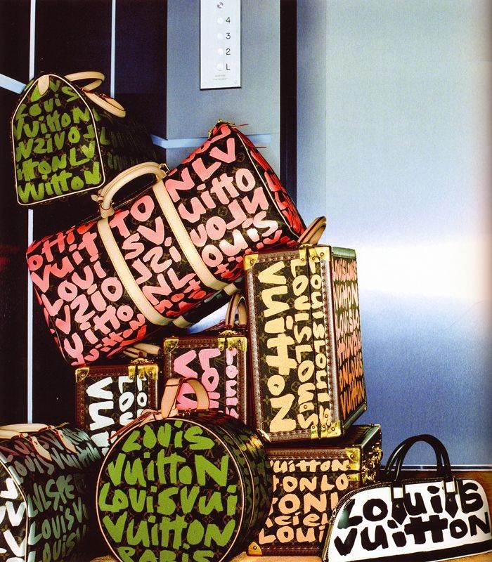 A collection of Louis Vuitton bags in various shapes, sizes, and colors, featuring the bold and playful graffiti-style monogram by artist Stephen Sprouse. The bags are stacked creatively against a modern backdrop, showcasing hues of pink, green, black, and brown with oversized, painted Louis Vuitton logos. The pieces include a mix of duffel bags, trunks, and round cases, each with a distinctive, eye-catching design.