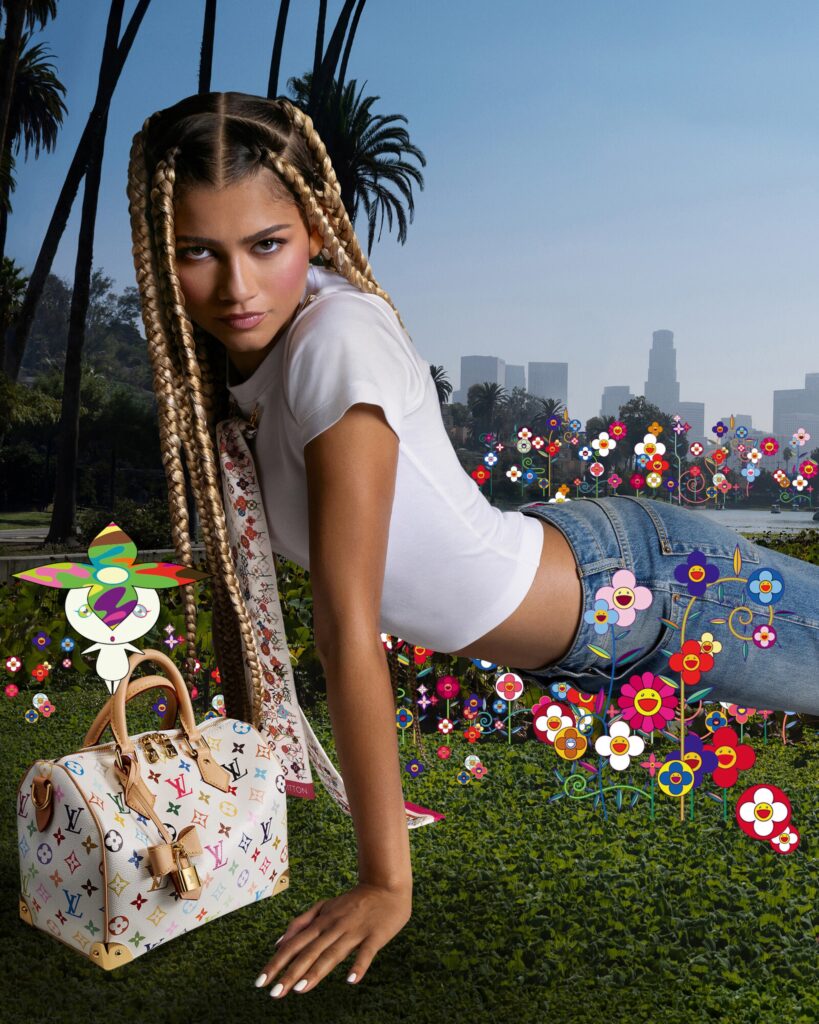 Zendaya posing in a vibrant outdoor setting with a Louis Vuitton x Takashi Murakami Multicolore Speedy bag. The scene features Murakami's signature colorful smiling flowers, blending high fashion with artistic surrealism against a Los Angeles backdrop.